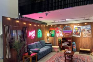 Custom Trade Show Booth— How Much Do They Cost? A vibrant 90's inspired living room set up for an experiential marketing event, featuring bold colors, patterned furniture, and nostalgic decor. The room includes a retro couch with geometric cushions, a vintage TV with a VHS player, a lava lamp, and posters of iconic 90's pop culture. The space is designed to immerse guests in a throwback atmosphere, with elements like cassette tapes, arcade games, and neon accents, evoking the energy of the 90's era.